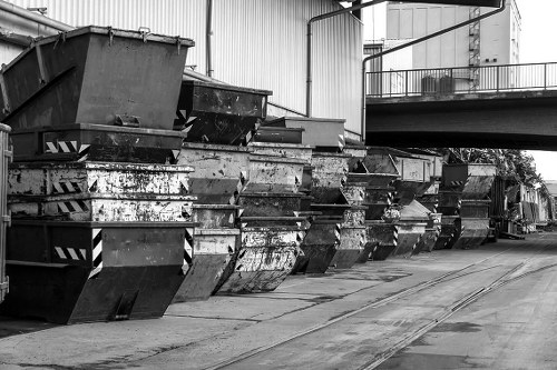A row of large, industrial metal skips lined up along a paved outdoor area, likely at a waste management or disposal site near commercial or warehouse premises. The skips are rectangular with open tops, showing signs of rust, dirt, and wear, indicating they are used for heavy waste collection. They are arranged in a straight line, with some slightly leaning or stacked behind others, suggesting active usage. The surface beneath is concrete with visible wear and tire marks, and a metal railing is visible on an elevated platform or balcony in the background. The surrounding environment includes the sides of large industrial buildings constructed from metal panels, with ventilation and piping visible overhead, creating an industrial atmosphere appropriate for a waste disposal service like Commercial Waste Euston serving the London area, potentially near the postal code region of NW1 or NW3. The overall scene is in natural outdoor lighting, emphasizing the utilitarian nature of the waste collection area.
