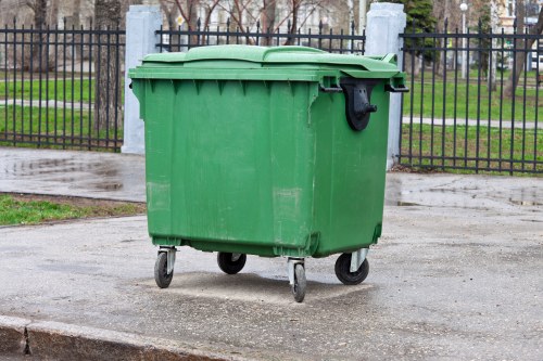 A green wheeled plastic waste bin with a closed lid, situated on a paved surface in an outdoor area near a black metal fence. The bin appears to be used for rubbish disposal, with a black handle on the side and small caster wheels at the bottom for mobility. Behind the fence, there are trees with bare branches and a grassy area, suggesting an urban or suburban setting in Euston or nearby. The pavement is slightly wet, possibly after rain, and the overall lighting is natural and diffuse. This scene represents typical rubbish collection infrastructure in a residential or public space, aligned with commercial waste management services provided by companies like Commercial Waste Euston.