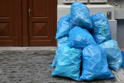 A collection of several large blue plastic rubbish bags filled with waste, piled on a paved sidewalk area in front of a brown wooden door. The bags appear full and tightly tied at the top, with some slightly bulging or leaning against each other. Surrounding the bags, the pavement is made of grey stone bricks, and part of a grey concrete structure is visible on the right side of the image. The setting is outdoors, likely on a residential or commercial property within central London, as suggested by the nearby door and urban environment. The scene captures a typical rubbish disposal arrangement, which Commercial Waste Euston might arrange for local businesses or tenants requiring waste collection services in the Euston area.