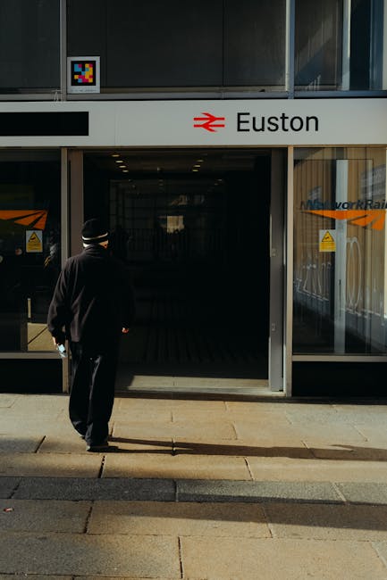 The image shows a section of the exterior of Euston station, featuring a large black signage panel displaying the white 'Euston' name alongside the iconic British rail and London Underground symbols. 